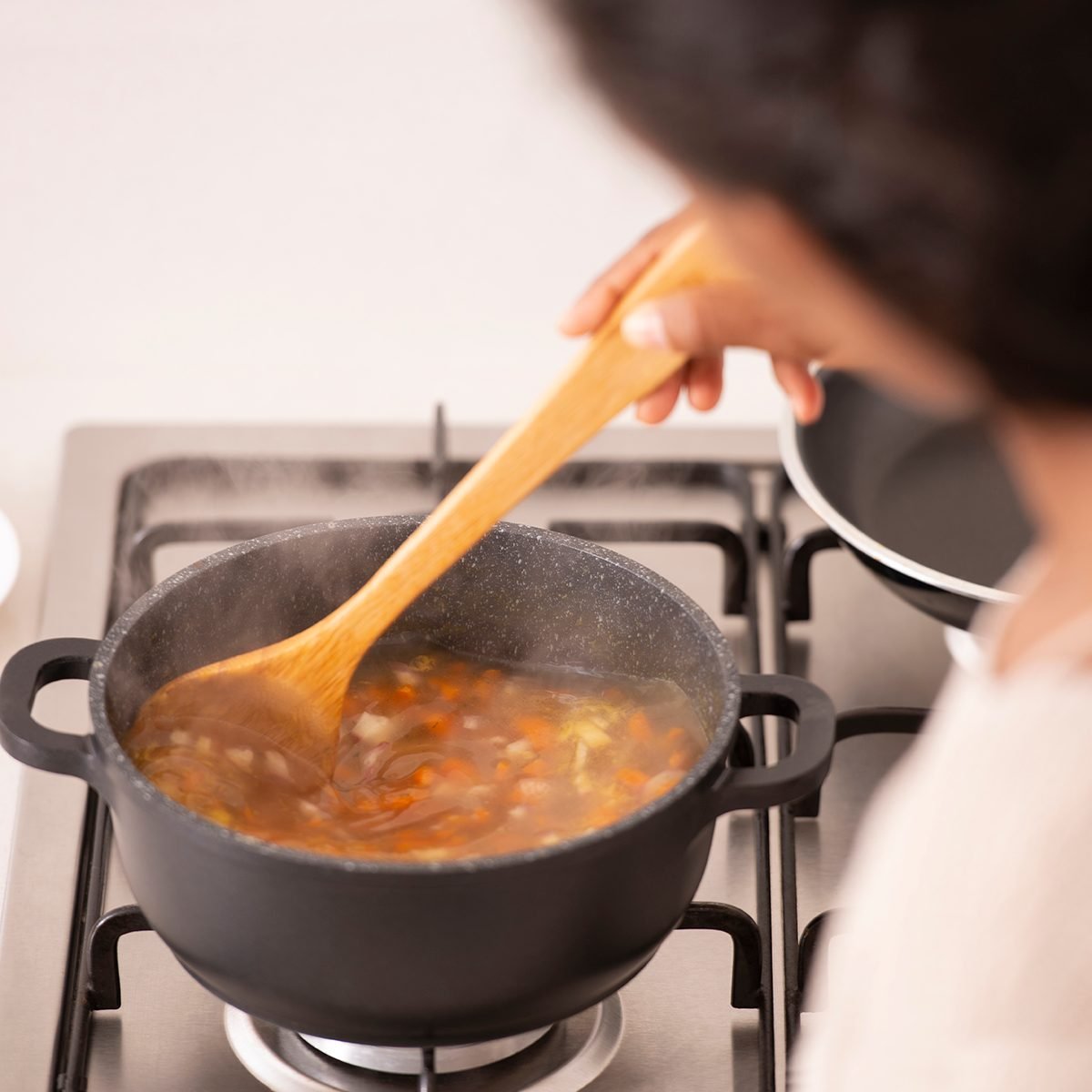 High angle, over the shoulder view of a woman stirring the boiling bouillon, using a wooden spoon. The woman cooking vegetable soup in a stock pot on a gas stove. A healthy meal, lifestyle and culinary concepts.