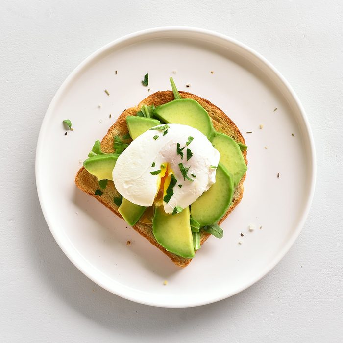 Poached eggs on toasted bread with avocado, rukola and herbs over white stone background with copy space. Top view, flat lay
