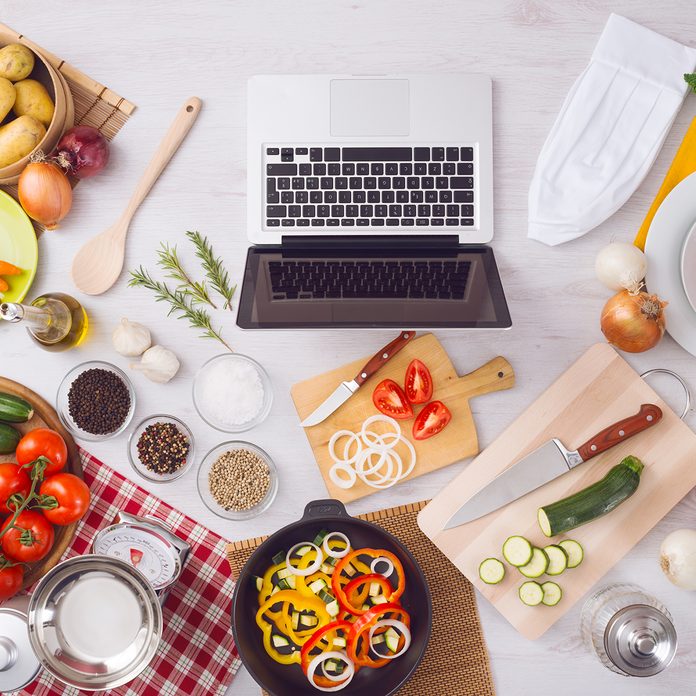 Home kitchen table top view with laptop, food ingredients, raw vegetables, kitchenware and utensils, top view