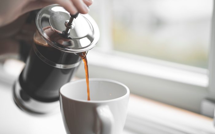 French style coffee press filled with hot coffee on a white window sill inside a house in Sweden. Close-up shot with selective focus of a person with tattoos pouring coffee into a cup.