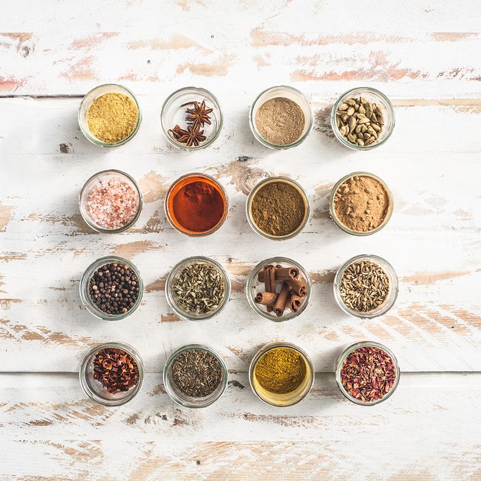 A flat lay display of various spices in small glass jars on a rustic wooden table top. Pink Himalayan salt, curry powder,cinnamon sticks, smoked paprika, cumin, peppercorns, chili, thyme, oregano, garlic powder, star anise, cardamon pods, pumpkin pie spice, fennel seeds, rose petals.