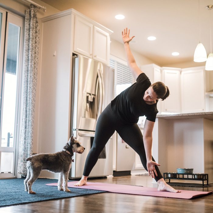 A mature adult woman does yoga and strength training exercises on a mat in her living room, her pet terrier dog keeping her company and trying to play.