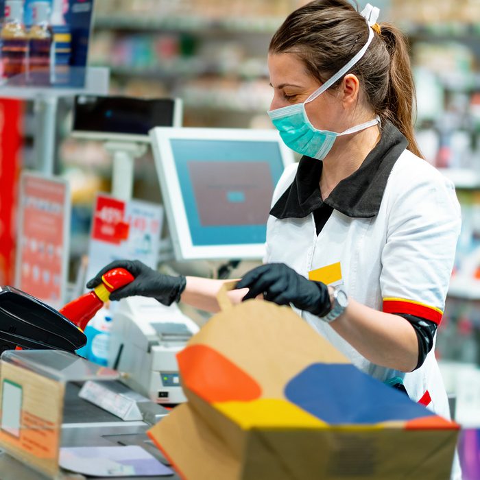 Woman cashier wearing protective face mask and gloves to prevent viruses, scanning disinfection products at the cash register and packing in paper bag
