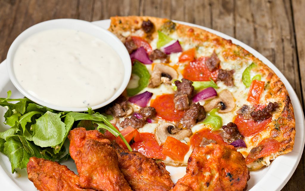 "A high angle extreme close up of a white paper plate with some spicy chicken wings , a generous portion of pizza,a small green salad and blue cheese dressing. Shot on a grungy old picnic table."
