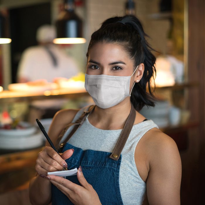 Portrait of a beautiful waitress working at a restaurant wearing a facemask while holding a notepad and looking at the camera â pandemic lifestyle concepts
