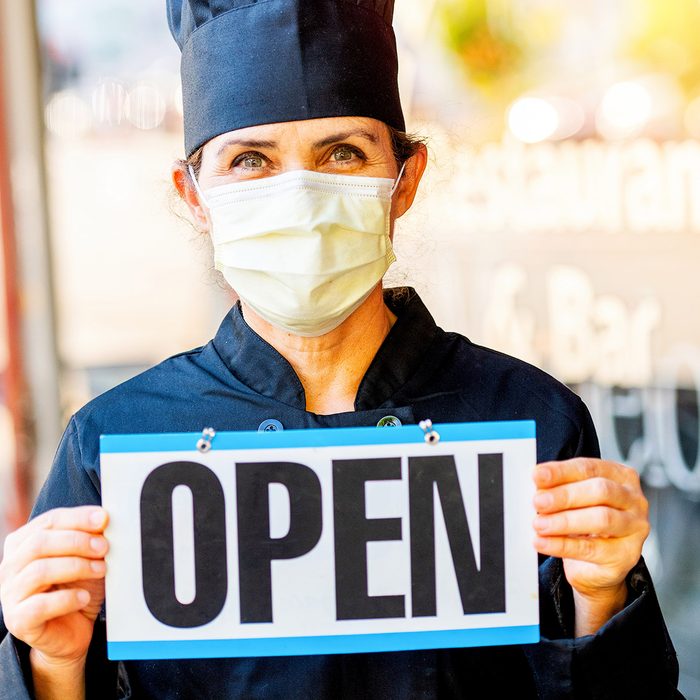 Female chef restaurant owner posing holding an Open Sign outside her restaurant wearing a mask because Covid-19