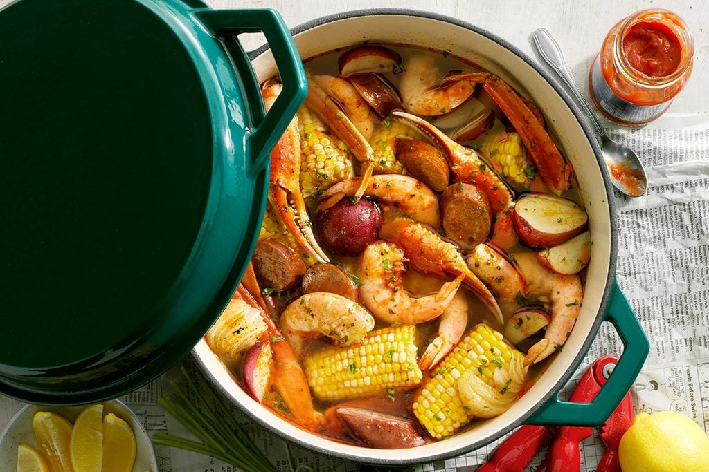 Low Country Boil; overhead camera angle; Wooden farm table surface; Taste of Home Cast Iron Dutch Oven; TOH enameled green 5 qt quart dutch oven with lid; mixed seafood; crab; shrimp; cilantro; sweet corn; potatoes; sausage; newspaper surface; oldbay tin; cocktail sauce; lemons; lemon wedges; Copy Space