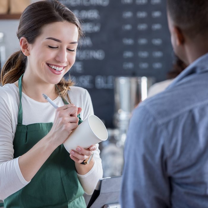 A smiling female coffee shop barista stands behind the checkout counter across from an unrecognizable patron. She looks down as she writes his order on a coffee cup.