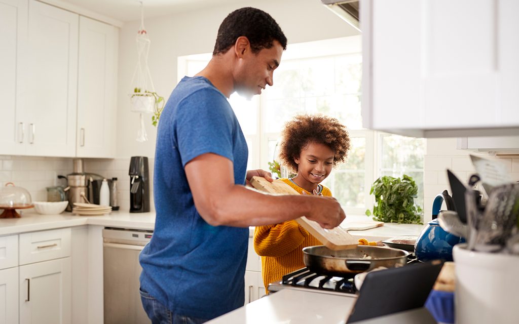 Pre-teen girl and her father standing at hob in the kitchen preparing food in a frying pan, following a recipe on a tablet computer, backlit
