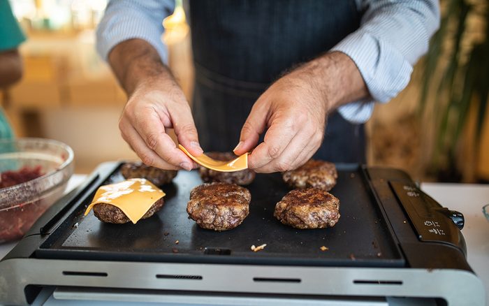Close up of human hands while unrecognizable man putting cheese on burgers while grilling them on electrical barbecue