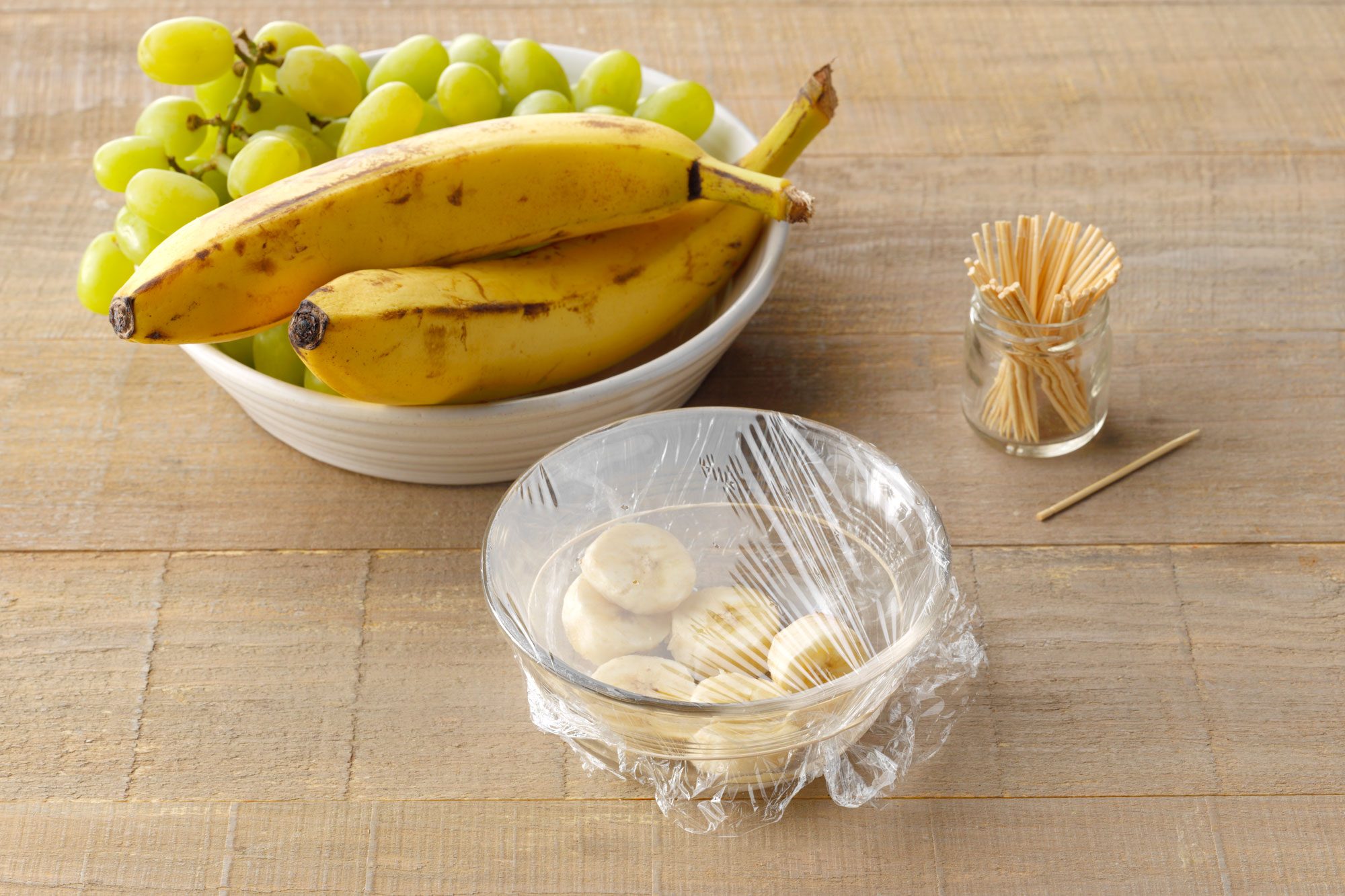 rotten fruit trap method. bowl of rotten banana slices in a bowl covered with plastic wrap that has had toothpick holes poked into it. wood surface background with bananas and grapes nearby.