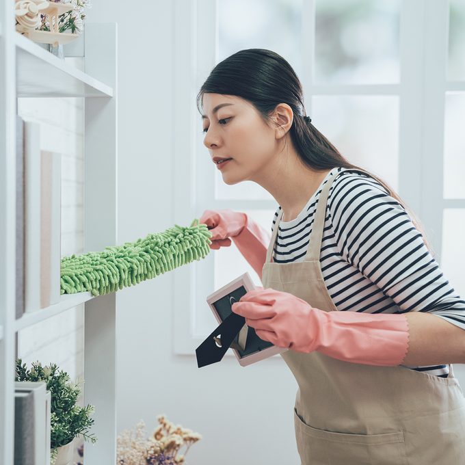 asian housekeeper in apron dusting the bookshelf by feather duster taking up the picture frame carefully cleaning in living room at home. young wife in rubber gloves doing housework.