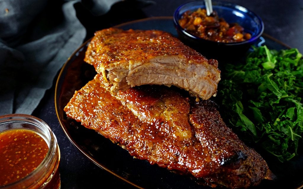 profile view of a platter of juicy oven ribs with a side of baked beans and greens