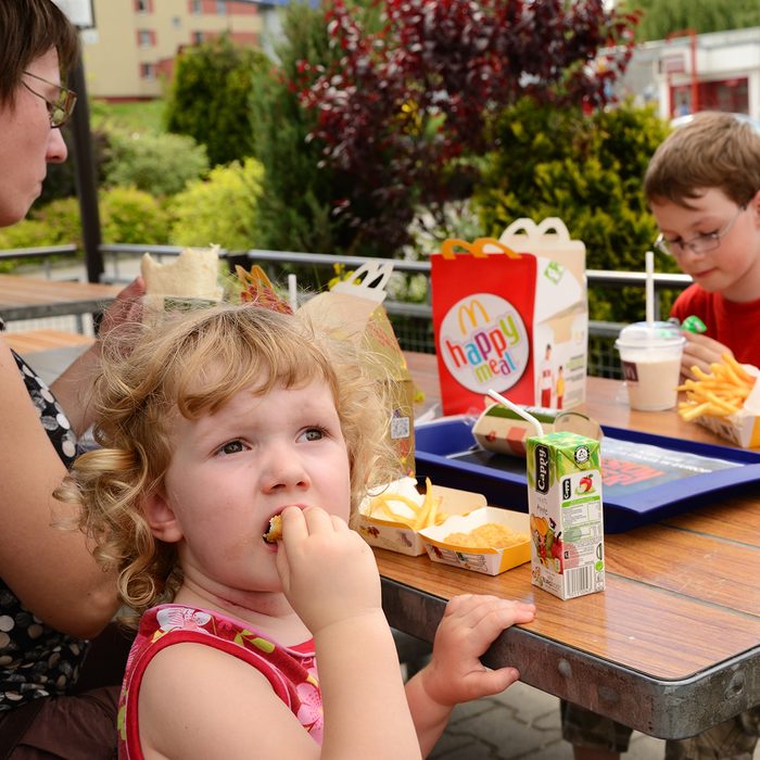 "Krakow, Poland - June 8th, 2012: Mother with two kids having a lunch in McDonald's, in the outdoor part of the restaurant. 3 years old blonde girl in the foreground, looking back and keeping a nugget in her mouth. Mother sitting on her left. 8 years old boy sitting opposite. Happy Meal box, some French fries, blue tray, milk shake, and a box with Cappy juice on the table.