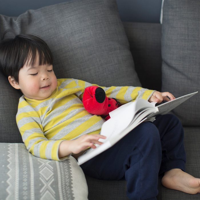 Asian toddler boy relaxing, reading on sofa.