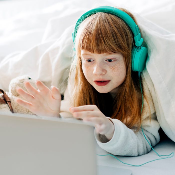 Young girl enjoying cozy time at home facetiming with grandparents via internet wearing headphones in bed on stay at home order during social distancing time due to health crisis virus outbreak