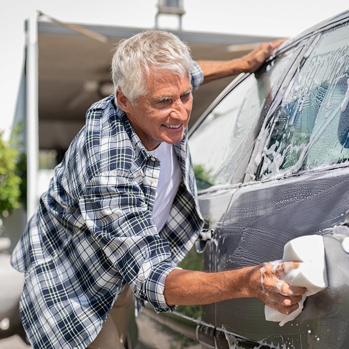 Happy senior man washing car with soap and foam. Old retired man cleaning automobile with sponge in a sunny day. Satisfied driver washing his gray car near the garage.