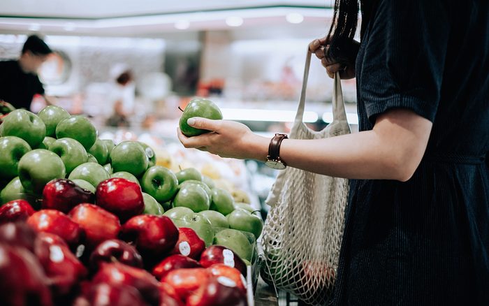 Cropped shot of young Asian woman shopping for fresh organic green apples in supermarket. She is shopping with a cotton mesh eco bag and carries a variety of fruits and vegetables. Zero waste concept