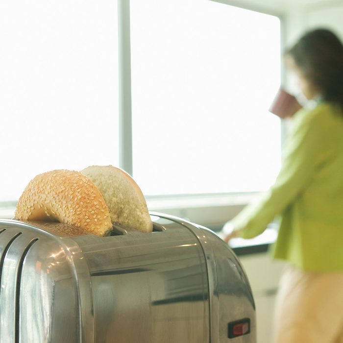 Woman in kitchen drinking coffee, bagels in toaster (focus on toaster)