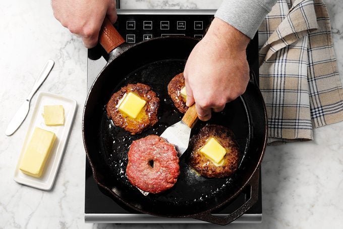 Easy Grilled Hamburgers; How to; marble surface; overhead camera angle; hands; burger; hamburger; burgers; hamburgers; butter; buttered; grilled; striped cloth; cast iron pan; cast iron skillet; juicy; frying; melted butter; butter knife; Step #7 Burger patties in pan with pads of butter on three fourth patty getting ready to be flipped; induction; prep; in process;