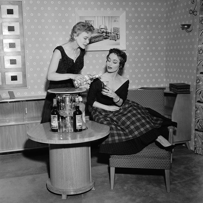 Models Jean Evans and Lynne Kennedy demostrating the push button cocktail cabinet at the British Furniture Exhibition at Earl's Court, London, 15th February 1954. The table has a concealed central section to store bottles and glasses which are revealed at the touch of a button. (Photo by Monty Fresco/Topical Press Agency/Getty Images)