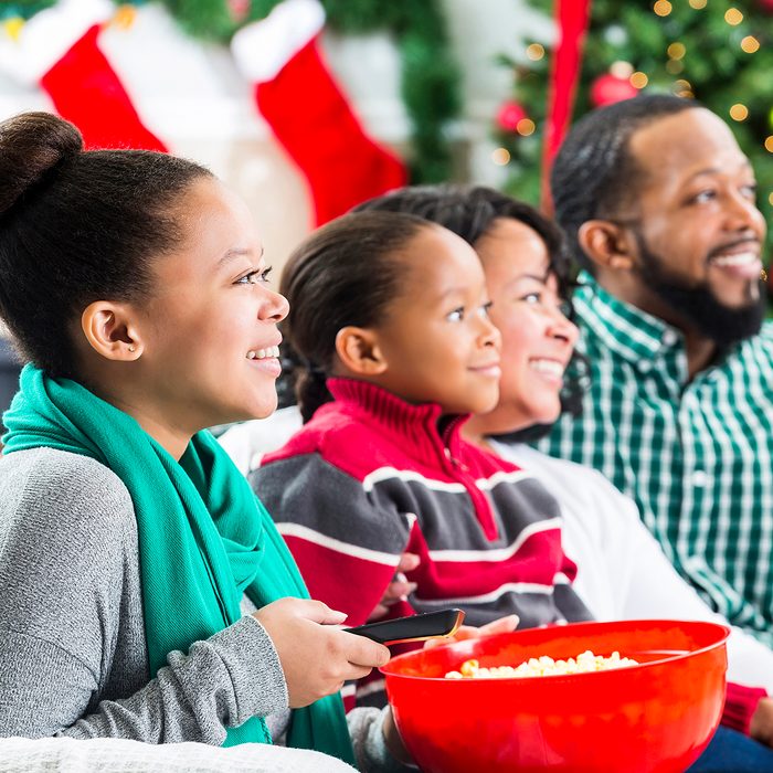 Happy African American couple and their teenage daughter and elementary age son watch a Christmas movie together during the Christmas season. The girl is holding a bowl of popcorn and the remote control. They are all smiling while watching the movie. A Christmas tree, stockings and a fireplace are in the background.