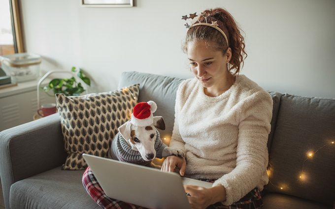 Young woman celebrating New Year at home with her dog. Dog is wearing costume, she is smiling and enjoy in holiday. Woman using laptop for online shopping