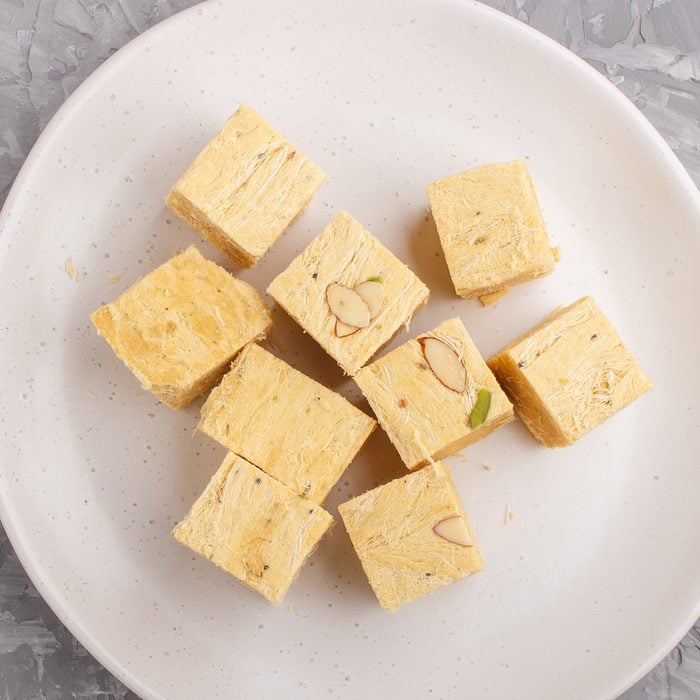 traditional indian candy soan papdi in white plate with almond and pistache on a gray concrete background. top view, flat lay, close up.