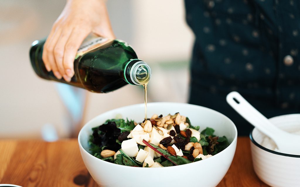 how to store olive oil Close-up view of a bowl of fresh green salad with mozzarella, mixed nuts and dry fruits. A woman’s hand was pouring olive oil into the salad.