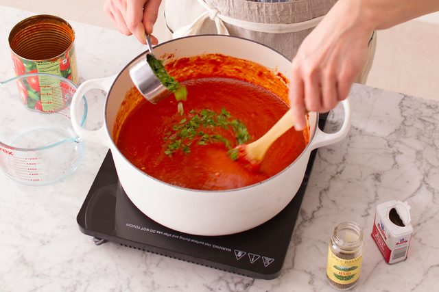 Person sprinkling in herbs into a pot on the stovetop with one hand and stirring sauce with a wooden spoon with their other