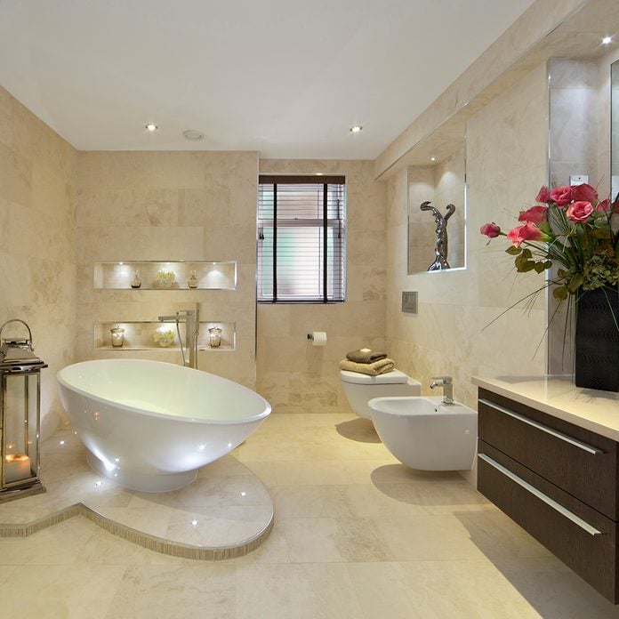 a modern bathroom in an expensive new home with a tear-drop shaped bath (full of water) sitting on a marble plinth. A bidet and WC are located near the window. A lantern with lit candles sits next to a large towel rail whilst a large bunch of red lilies sit on the cabinet to the right of the picture.