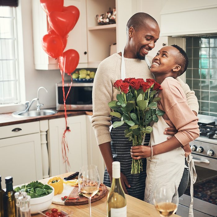 Cropped shot of an affectionate young couple posing with a bunch of flowers in their kitchen on their anniversary