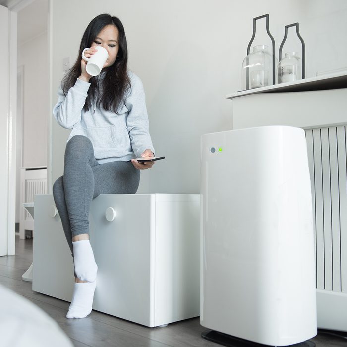 A Young Asian Woman Controls The Power Of Her Smart Air Unit From Her Mobile Phone As She Sits On A Storage Bench And Enjoys A Cup Of Tea In The Bedroom