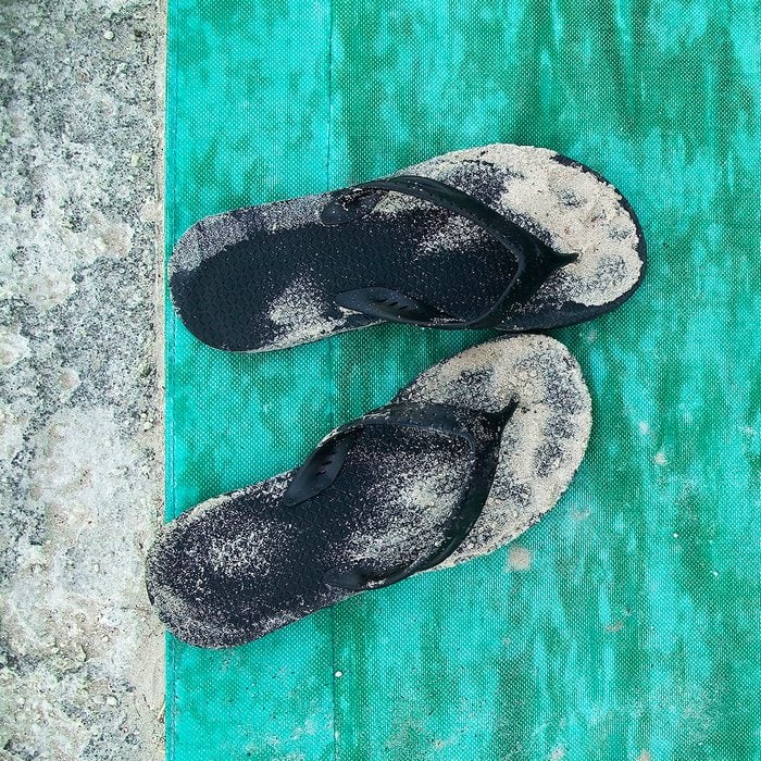High Angle Close Up Of Sandy Black Flip Flops On Turquoise Floor Mat.