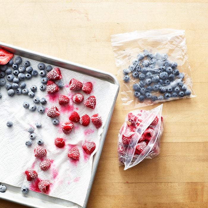 how to make frozen drinks Overhead of freezing berry preparation, rasperries, blueberries and strawberries sit frozen on a paper towel on a silver baking sheet with plastic bags holding more frozen berries sitting next to the tray on a wooden surface.