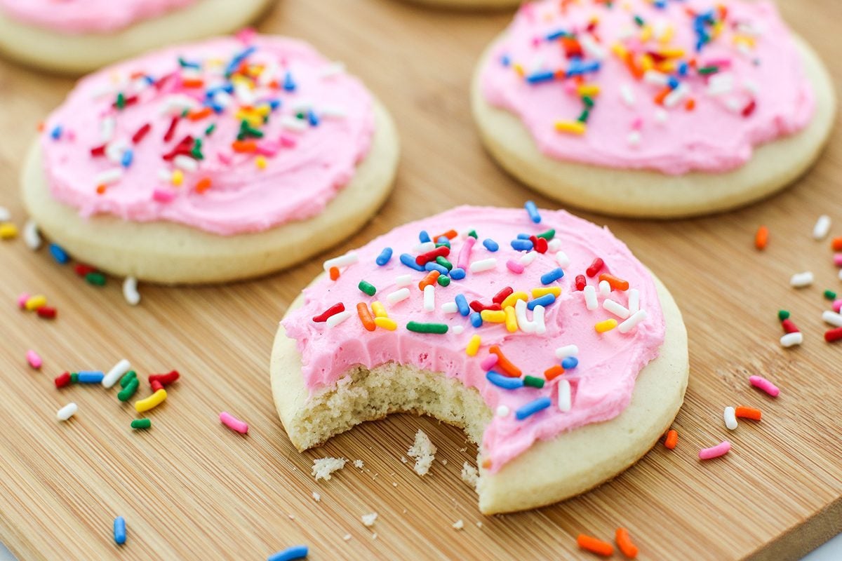 Lofthouse Cookies with pink frosting and sprinkles on a wooden cutting board, one cookie has a bite taken out of it