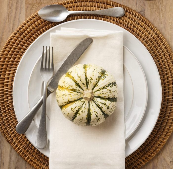 Festive Fall Thanksgiving table setting place setting home decorations with white china plates dishes, silverware, linen cloth napkin, placemat, white wine in wine glass, natural miniature pumpkins and ornamental squash on rustic wooden tab