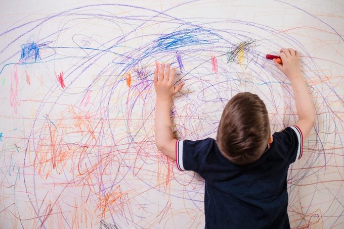 The Child Draws On The Wall With A Crayon. The Boy Is Engaged In Creativity At Home