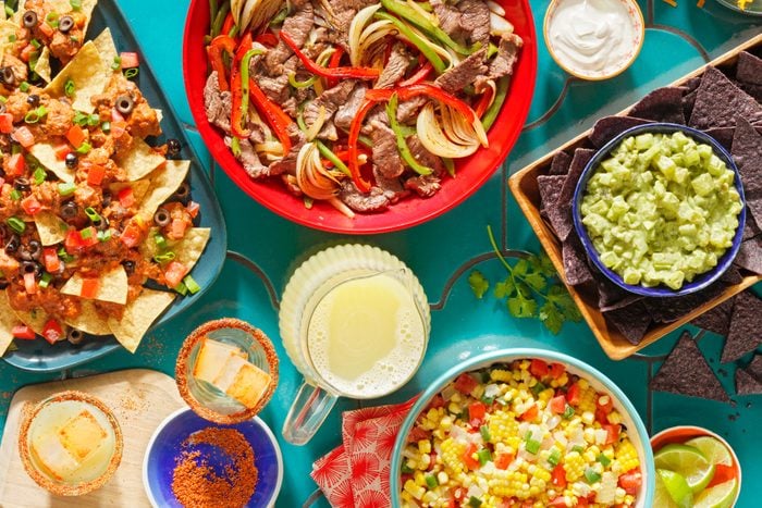 overhead view of colorful tex-mex food, nachos, fajitas, chips and guacamole, corn salsa, and a pitcher of margaritas on a teal tiled table