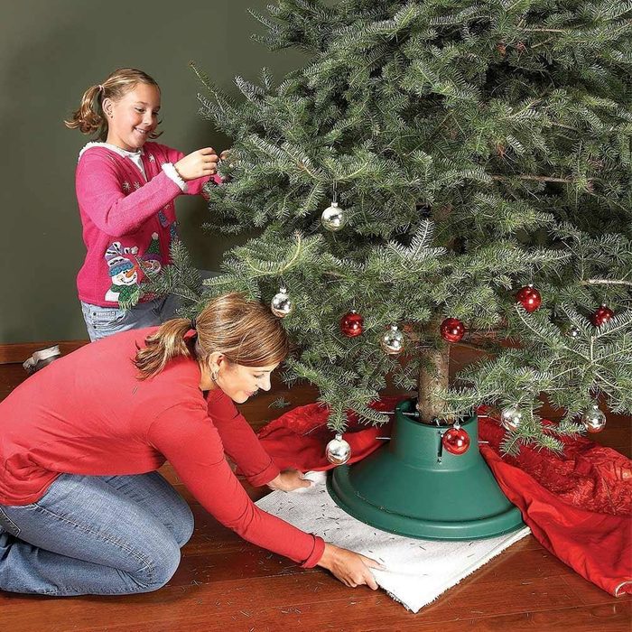 a woman adjusting the base of a christmas tree while a child adds ornaments to the tree