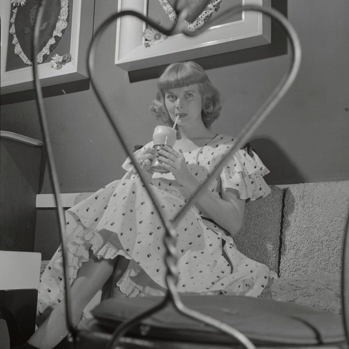 Looking demure as an old-fashioned valentine, Dorothy Abbott, drinking an ice-cream soda, is seen through the frame of a wire-back chair. Her pink cotton dress has ruffles at the shoulder and a black grosgrain ribbon ties around her waist and extends to hemline.
