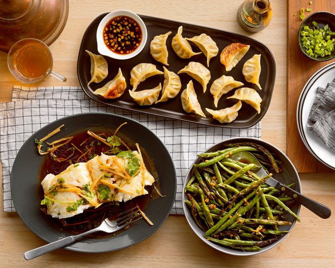 lunar new years dishes on a wooden table; homemade pork dumplings, ginger scallion and soy steamed fish, and sesame soy string beans