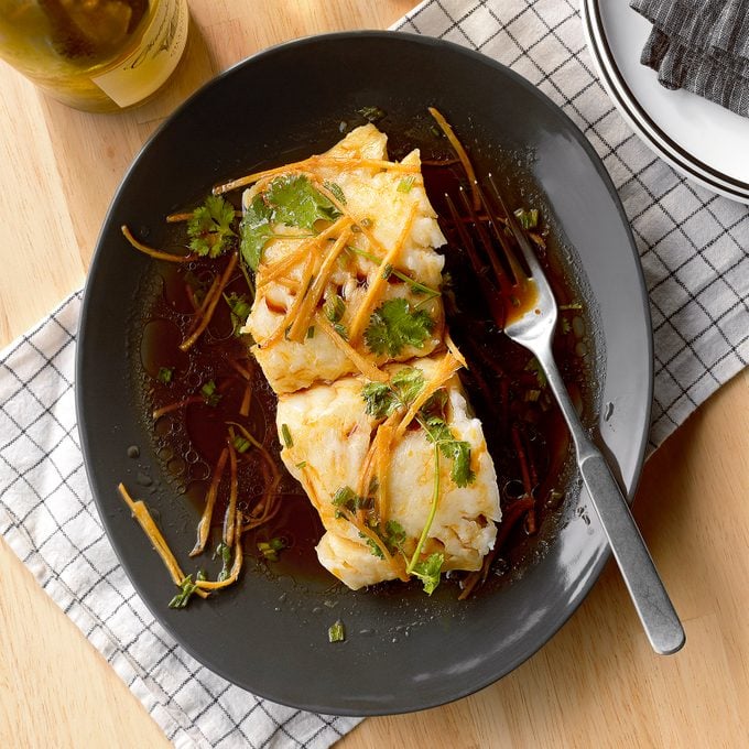 Ginger Scallion And Soy Steamed Fish on a dark plate with a fork on a light wood table