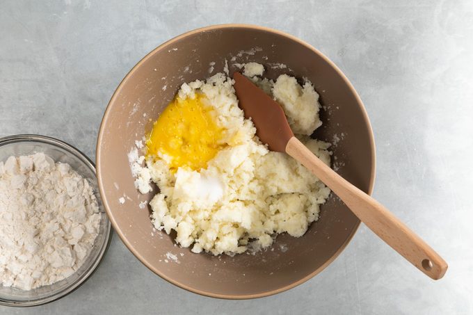 mashed potatoes in a large bowl with a raw egg and salt added, ready to be mixed. bowl of flour sits nearby.