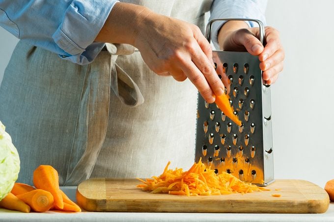 Woman Grates Fresh Carrots For Cooking Salad Or For Salting Cabbage Natural Background. Vegetarian Food Concept.