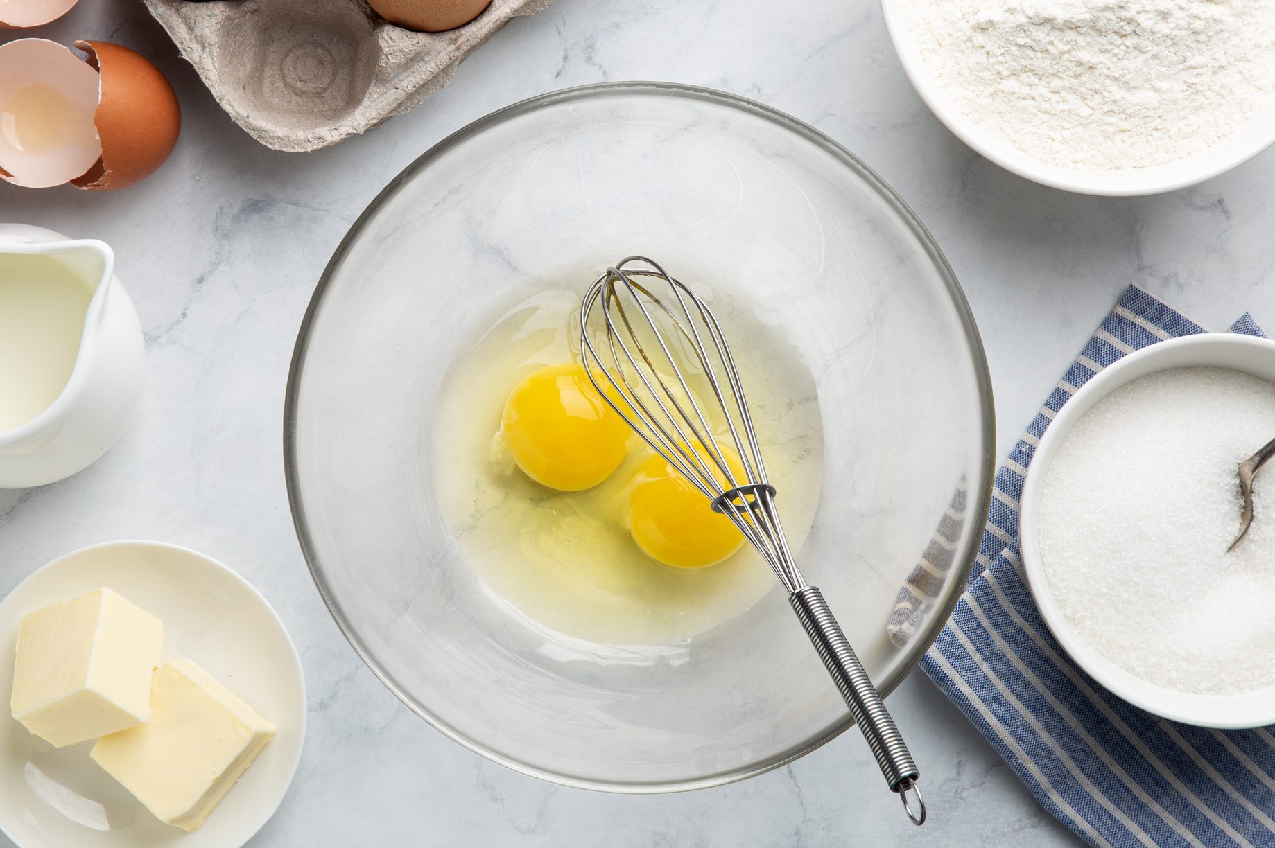 raw eggs in a glass bowl with a whisk surrounded by other baking ingredients