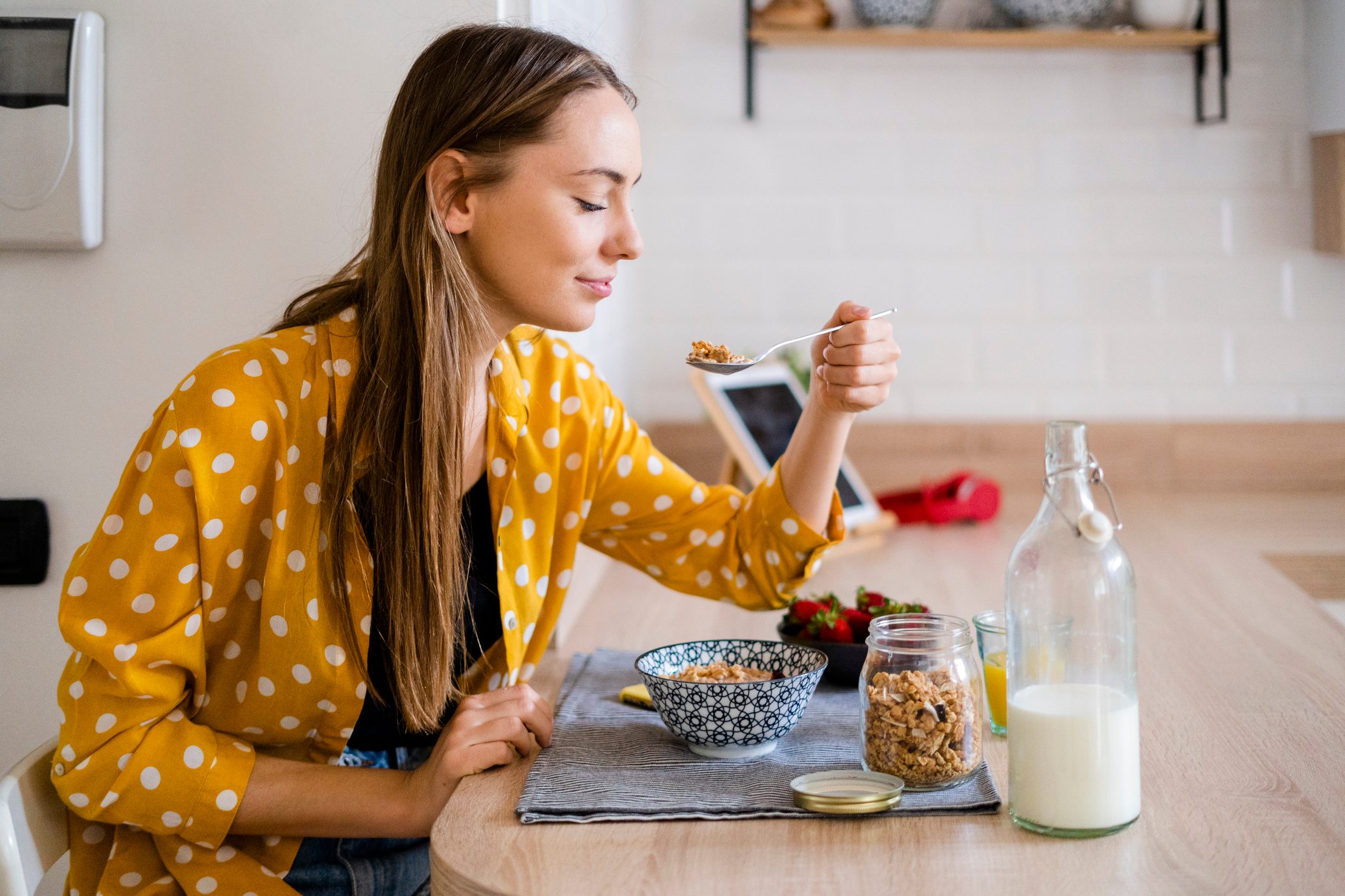 Young woman enjoying breakfast in kitchen at the kitchen table, eating a bowl of cereal on a placemat with a bottle of milk next to it