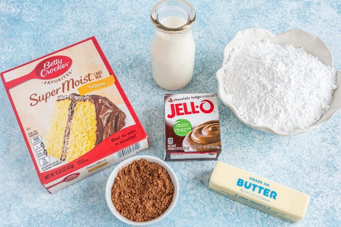 ingredients for making cake on a blue counter top including a cake mix box, jello pudding box, jar of milk, flour, and butter