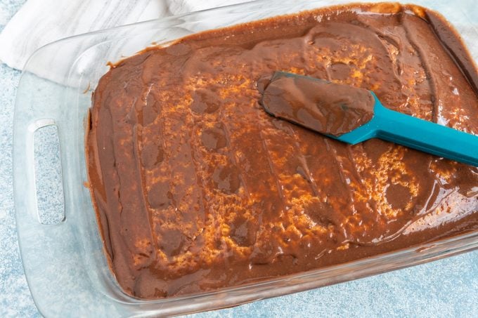 cake in a glass pan with a thin layer of chocolate pudding covering the whole cake and a blue rubber spatula resting on top