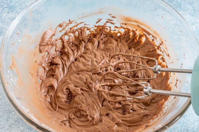 chocolate frosting in a large clear glass bowl with electric beaters resting on the side